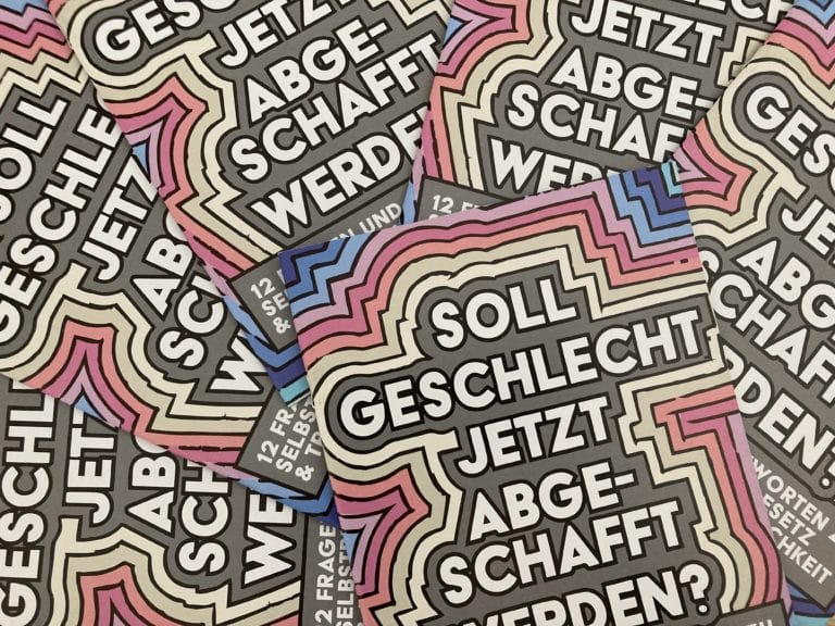 6 Exemplare der Broschüre liegen auf einem Tisch. Das Titelbild ist nur bei einer Broschüre ganz zu sehen. Auf dem Titelbild steht in großer Schrift: „Soll Geschlecht jetzt abgeschafft werden?“ Darunter steht kleiner Schrift: 12 Antworten auf Fragen zum Thema Selbstbestimmungsgesetz und Trans*geschlechtlichkeit." Die Schrift ist in einem grauen Kasten. Dieser Kasten hat bunte Umrandungen: Erst kommt ein gelber Rand, dann ein oranger, dann ein pinker, dann ein blauer. Dadurch entsteht ein Regenbogen um den Titel der Broschüre.