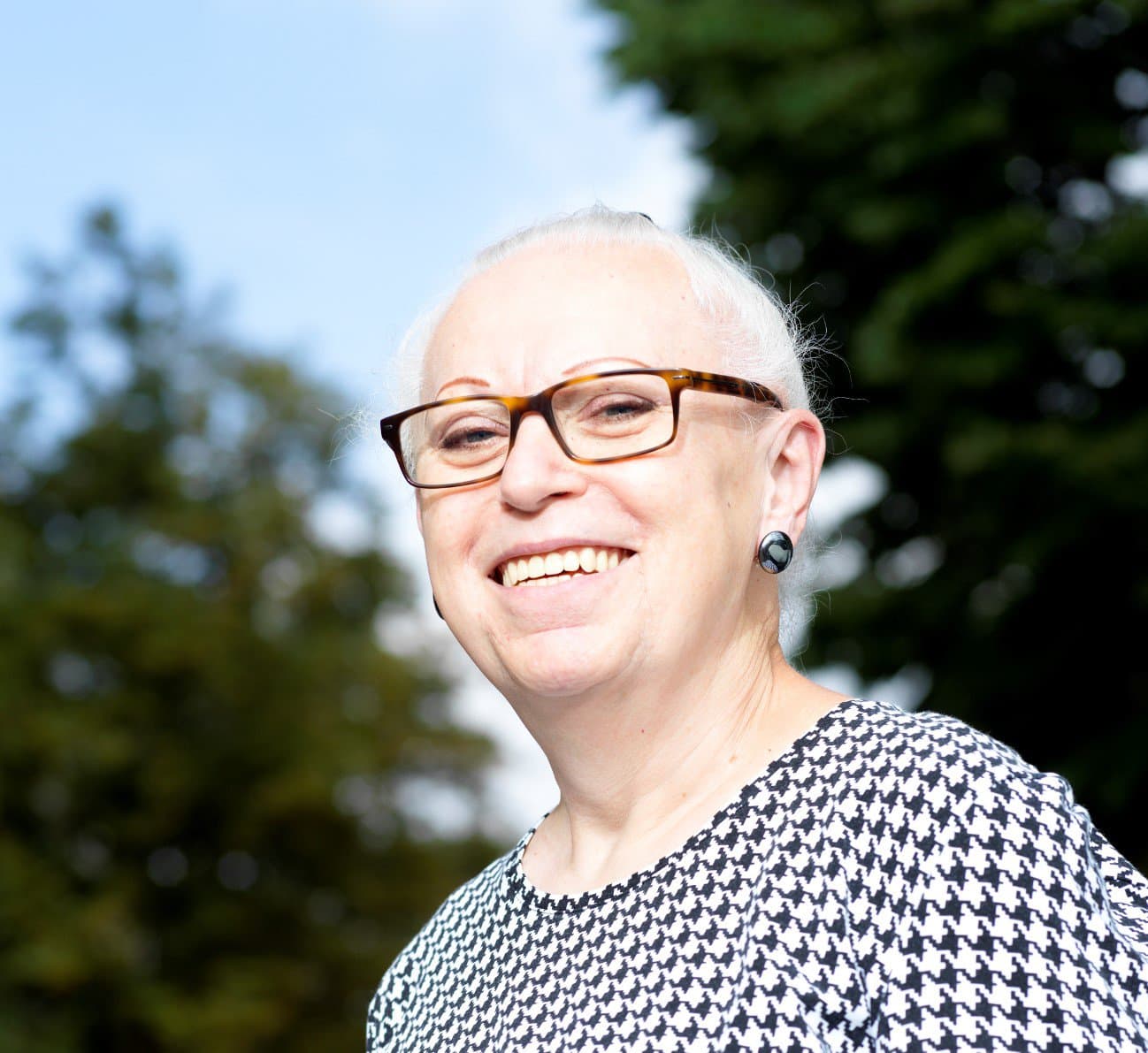 Portrait von Nora lächelt vor einem verschwommenen Hintergrund mit Baumwipfeln in die Kamera. Sie trägt eine Brille und ein schwarz-weiß kariertes Oberteil.