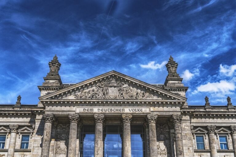 Auf dem Foto ist der Deutsche Bundestag zu sehen. Das Foto zeigt das Gebäude in Nahaufnahme. Der Schriftzug "Dem Deutschen Volke" ist auf dem Gebäude zu lesen. Über dem Dach ist ein blauer Himmel zu sehen, der von vielen weißen und grauen Wolken bedeckt ist.