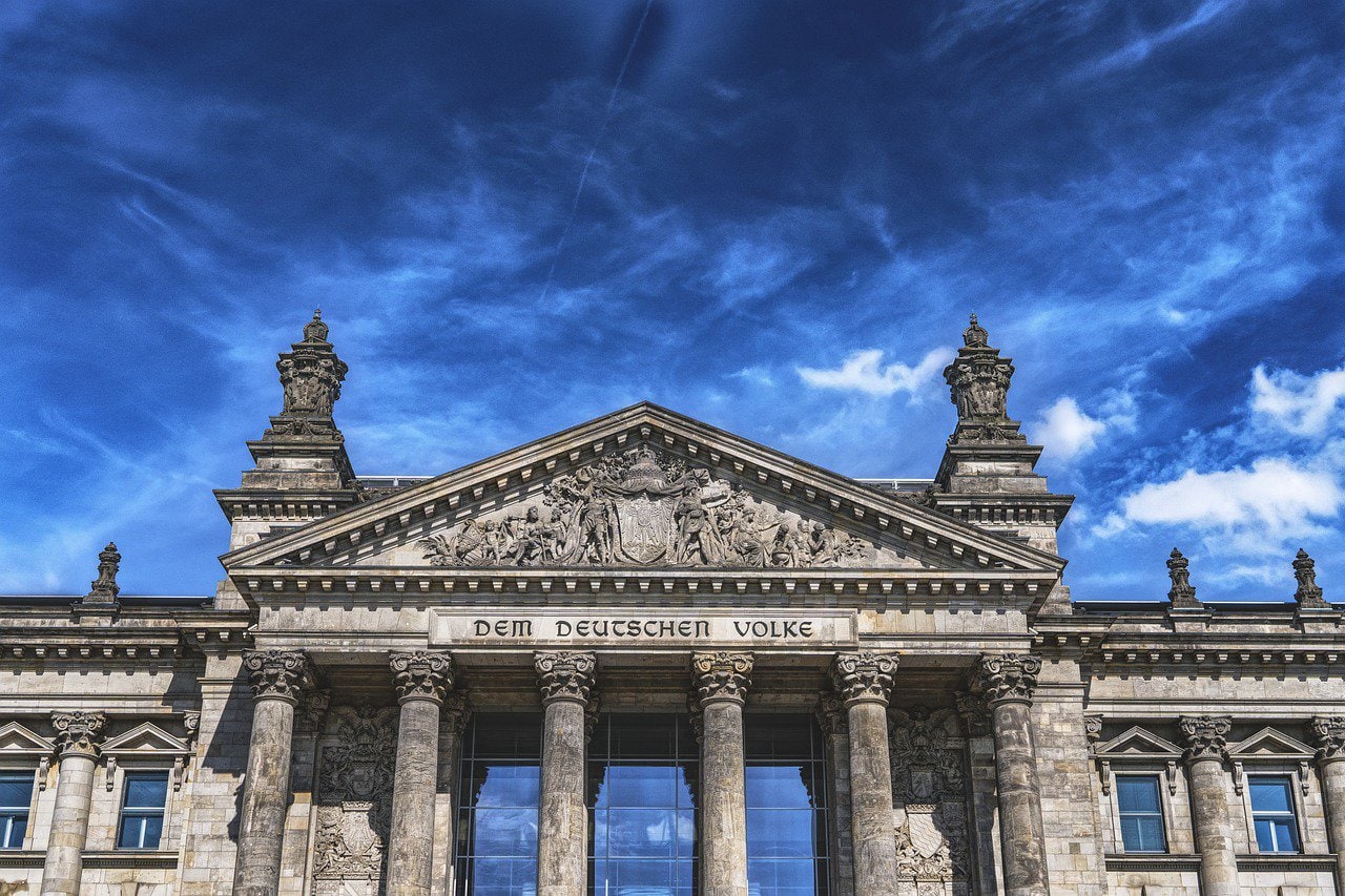 Auf dem Foto ist der Deutsche Bundestag zu sehen. Das Foto zeigt das Gebäude in Nahaufnahme. Der Schriftzug "Dem Deutschen Volke" ist auf dem Gebäude zu lesen. Über dem Dach ist ein blauer Himmel zu sehen, der von vielen weißen und grauen Wolken bedeckt ist.